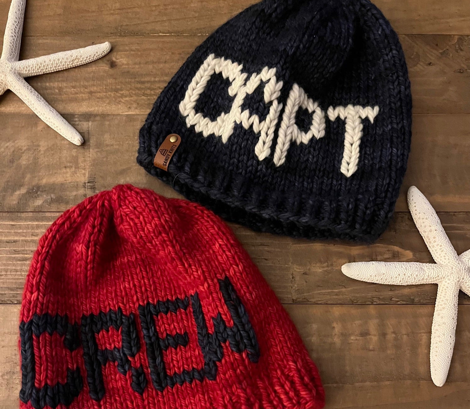 Two knit hats, one red and one black with white text, on a wooden surface with starfish.