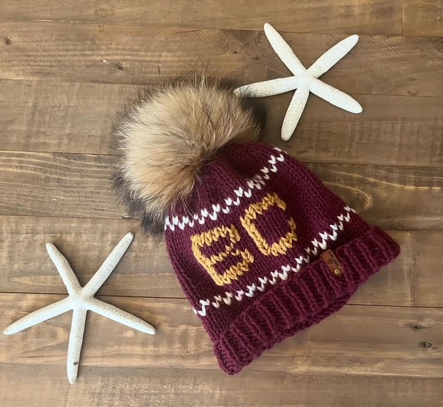 Maroon knit beanie with fur pom-pom and 'BC' lettering on a wooden surface with starfish.