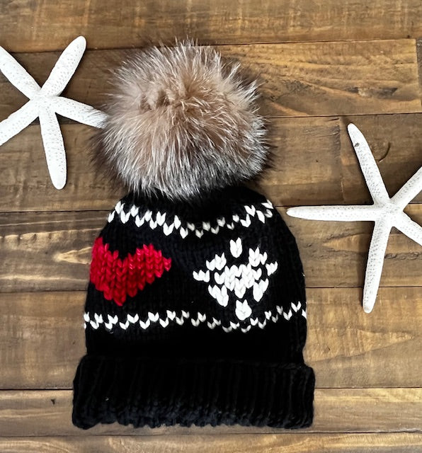 Black knit beanie with a large pom-pom and red heart design on a wooden surface.