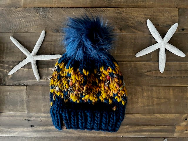 Colorful knit beanie with a blue pom-pom on a wooden surface with starfish decorations.