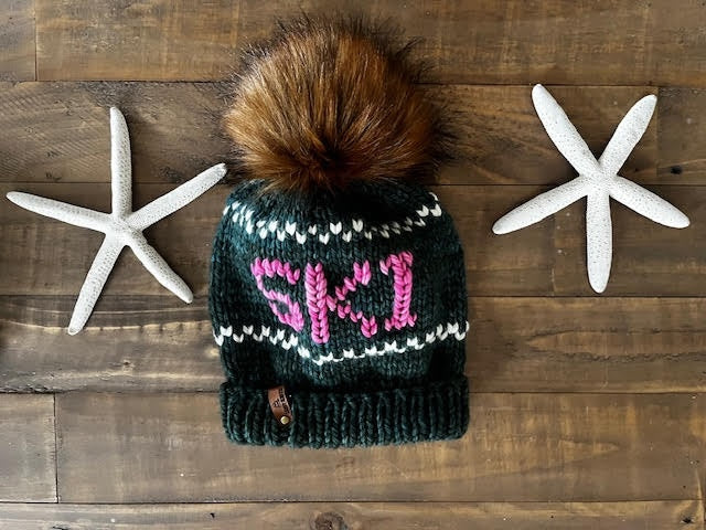 Knitted beanie with pom-pom and 'SKI' text on a wooden surface with starfish